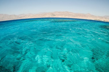 Kızıl deniz, berrak gökyüzünün altında çarpıcı turkuaz suları sergiliyor. Engebeli dağlar uzak bir zemin oluşturarak sakin okyanusla kontrastı güçlendirir. Parlak gün ışığı sahneyi aydınlatıyor