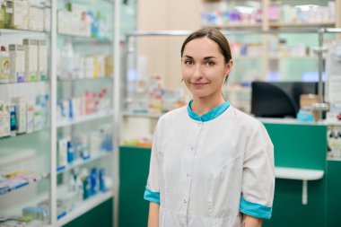 Kahverengi saçlı ve beyaz önlüklü açık tenli genç bayan eczacı. Parlak eczanede güvenle ayakta duruyor. Arka planda bulanık ilaç rafları davetkar bir atmosfer yaratır.