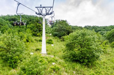 Çelik teleferik kulesi, sık ağaçlarla çevrili yeşil dağ yamacında duruyor. Bulutlu gökyüzü, canlı yeşillikleri vurgulayarak nötr bir zemin oluşturur. Yokuş yukarı bakış açısı yüksekliği ve eğimi vurgular
