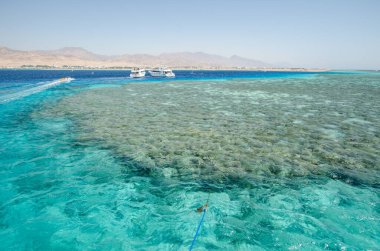 Açık gökyüzünün altında Kızıl Denizler 'in nefes kesen canlı mercan resiflerinin çarpıcı hava perspektifi. Yatlar yakınlarda yüzer, uzak ufukta dağlık bir manzara, doğal güzelliği ve dinginliği gösterir.