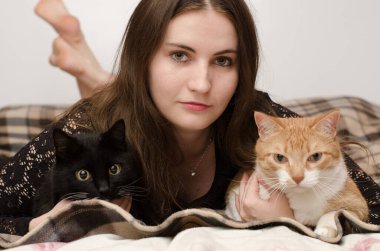 Young woman with brown hair lies on bed with black and ginger cats. Cozy setting with checkered blanket and neutral background. Calm expressions create warm atmosphere clipart