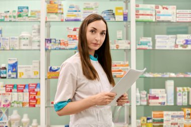 Kahverengi saçlı, genç, yetişkin bir kadın eczacı eczanede güvenle duruyor. İyi düzenlenmiş ilaç raflarıyla çevrili bir panosu var. Parlak ışıklandırma profesyonel atmosferi vurgular
