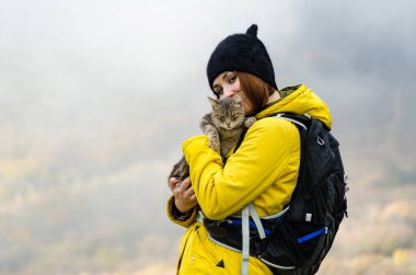 Uzun saçlı, sarı ceketli, siyah şapkalı, sırt çantalı, sisli dağda kedi tutan genç bir kadın. Hafif ışıklandırma, sis dingin bir hava yaratır. Bulanık arkaplanda sonbahar renkleri
