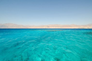 Kristal berraklığında turkuaz sular, bulutsuz gökyüzünün altındaki kurak dağlarla Kızıl Deniz boyunca uzanıyor. Huzurlu deniz manzarası deniz ve karanın canlı kontrastını sunar