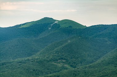 Bulutlu gökyüzü altındaki yemyeşil dağlar ve vadilerin geniş manzarası. Yoğun ormanlarla kaplı, dağınık yolları olan yuvarlanan bir arazi. Yumuşak ışık huzurlu bir atmosfer yaratır.