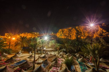 Gece gökyüzünün altındaki çöl tatil köyünde, kayalık bir zemin var. Hasır şemsiyeler ve şezlonglar düzenli bir şekilde dizilmiş, yumuşak, sıcak ışıklandırmalı palmiye ağaçları