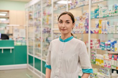 Kahverengi saçlı genç bir kadın eczanede beyaz önlük giyiyor. Etrafını renkli ilaç kutularıyla dolu raflar sarmış. Parlak ışıklandırma temiz ve düzenli atmosferi geliştirir.