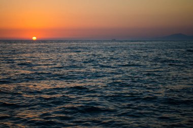 Turuncu günbatımı, sakin okyanus sularının üzerine sıcak bir ışık saçar. Uzak dağların silueti canlı gökyüzüne karşı huzurlu ve manzaralı bir manzara sunuyor. Büyüleyici doğal güzellik.