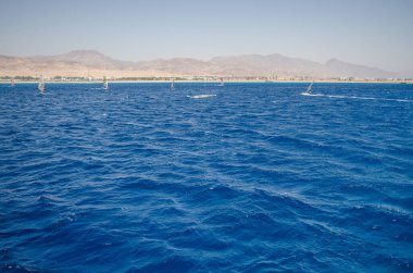 Kızıl Deniz 'in bulutsuz gökyüzü altında canlı mavi dalgalarla geniş bir manzarası var. Uzak dağlar ufuk çizgisinde çarpıcı bir kontrastlama yaratıyor. Huzurlu deniz manzarası sakinliği çağrıştırıyor
