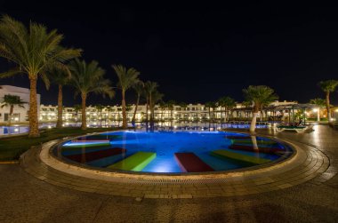 Renkli şezlonglarla aydınlatılmış havuzun olduğu sakin bir tatil yeri sahnesi. Palmiye ağaçları açık gece gökyüzüne karşı havuz kenarında. Sıcak ışıklandırma, mimariye tesis oldu