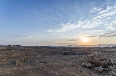 Sabah ışığında geniş bir çöl manzarası. Yumuşak güneş ışığı kurak arazide yumuşak gölgeler bırakır. Bulutlu gökyüzü arka plandaki kumlu renkleri tamamlıyor