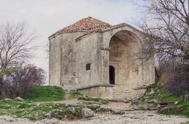 Kırım 'daki kayalık arazide bulunan yıpranmış bir kilise. Çorak ağaçlarla çevrili yapılar, yaş ve tarih belirtileri gösteriyor. Bulutlu gökyüzü yumuşak, dağınık ışık yayar, sakin bir atmosfer yaratır.