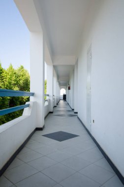 Beyaz duvarları ve parlak doğal ışık altında döşenmiş uzun otel koridoru. Koridor uzak kapıya çıkar, yemyeşil ağaçlar mavi gökyüzünün yanında görünür.