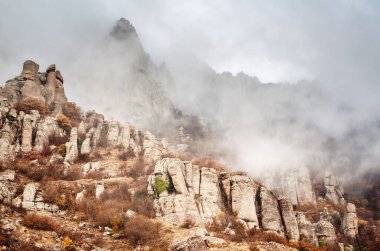 Kırım 'daki Demerdzhi dağının sisinin içinden engebeli kaya sütunları yükseliyor. Yumuşak, dağınık ışıklandırma eşsiz jeolojik özellikleri vurgular. Topraksı tonlar ve bulutlu gökyüzü ürkütücü, mistik bir hava yaratır.