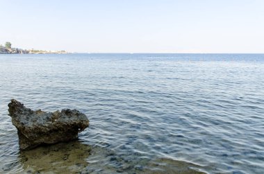 Işıl ışıl mavi denizin berrak gökyüzüne kavuştuğu sakin sahil manzarası. Ön planda sert kayalar ve doku eklenirken, uzak ufuk barışçıl geri çekilme ipuçları veriyor.