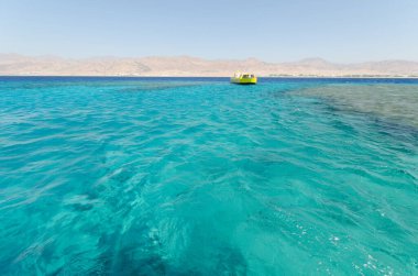 Parlak sarı tekne, açık gökyüzünün altında turkuaz kızıl denizle çelişiyor. Arka plandaki çöl dağları egzotik yerleri geliştirir. Güneş ışığı sakin deniz yüzeyine yansıyor