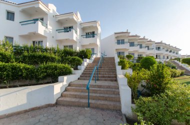 Beyaz cepheli ve simetrik tasarımlı zarif tatil köyleri binaları. Canlı yeşil bahçeler taş girişlere giden yollar. Açık mavi gökyüzü dingin atmosferi arttırır