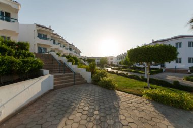 Tatil köyünün girişinde ağaçlar ve yeşil çalılarla sıralanmış basamaklar görülüyor. Güneş batışı sıcak, davetkar bir parıltı yaratır. Yol kırsal cazibeyi artıran desenli taş işçiliğine sahip.