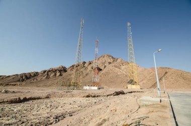 Uzun iletişim kuleleri, açık mavi gökyüzünün altında çorak ve kayalık arazinin ortasında duruyor. Güneş ışığı, engebeli arazinin üzerine, ıssız bir çevreyi aydınlatan farklı gölgeler bırakır.