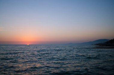 Sıcak, değişken günbatımı gökyüzü altında sakin bir okyanus manzarası. Güneş kısmen ufkun altına batıyor, turuncu parlıyor. Yumuşak dalgalı ve geniş manzaralı huzurlu bir atmosfer.