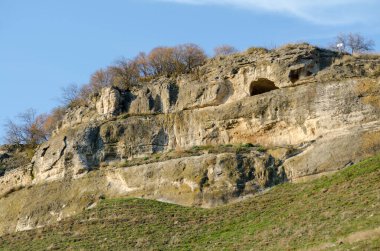 Kırım 'daki güneşli uçurum yüzeyi engebeli kayaları ve mağara açıklarını gösteriyor. Zirvedeki seyrek bitki örtüsü doğal manzaraya doku ekler. Açık gökyüzü dingin güzellikleri vurguluyor