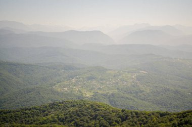 Puslu gökyüzünün altındaki yemyeşil ormanların ve görkemli dağların geniş manzarası. Uzak tepeler ufka karışıp huzur dolu manzaralar yaratıyor. Yumuşak, doğal ışıklandırma huzurlu atmosferi geliştirir.