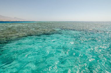 Kızıl Deniz 'in turkuaz suları parlak güneş ışığı altında parıldıyor. Dalgalanan yüzey uzak dağlarla buluşarak sakin ve nefes kesici deniz manzarası yaratıyor. Açık gökyüzü canlı manzarayı güçlendirir