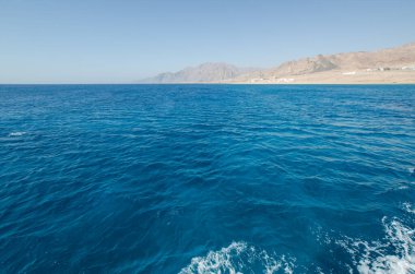 Kızıl Deniz 'in berrak mavi suları parlak gün ışığında uzak kayalık dağlara doğru uzanıyor. Huzurlu ufuk, deniz ve gökyüzünü harmanlayarak sakin deniz kenarı manzarası yaratır.