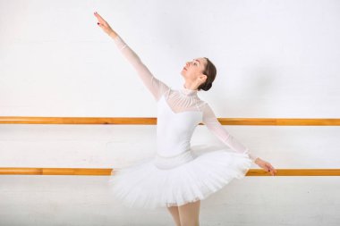 Açık kahverengi saçlı, bale barında zarif poz veren genç bir bayan balerin. Beyaz tayt ve etek yumuşak ışıklandırılmış stüdyo geçmişine karşı sakin ifadeyi artırır