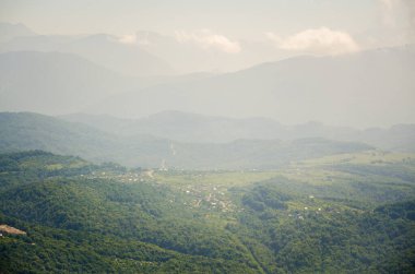 Berrak mavi gökyüzünün altındaki yemyeşil dağların geniş manzarası. Yuvarlanan tepeler ve vadiler, yumuşak gün ışığıyla çevrili olan mesafeye kadar uzanıyor. Yeşillik manzaralı küçük köyler