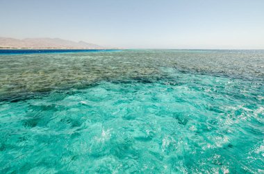 Kızıl Deniz 'in turkuaz suları parlak güneş ışığı altında parıldıyor. Dalgalanan yüzey uzak dağlarla buluşarak sakin ve nefes kesici deniz manzarası yaratıyor. Açık gökyüzü canlı manzarayı güçlendirir