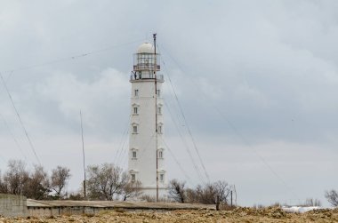 Majestic deniz feneri bulutlu gökyüzünün altında kayalık sahilde dimdik ayakta duruyor. Yakınlarda seyrek bitki örtüsü ve anten, sakin ve izole deniz manzarası yaratıyor. Yumuşak ışıklandırma yapıların detaylarını vurgular