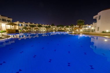 Geniş tatil köyü havuzu, sakin gece gökyüzünün altında mavi parlar. Etrafını saran beyaz binalar, sakin atmosferi arttırıyor. Rahatlamak ve düşünmek için mükemmel.