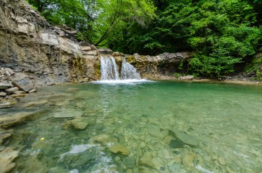 Küçük şelaleler yemyeşil ormanlarla çevrili kristal berraklığında havuza dökülür. Taşlar ve kayalar şeffaf suyun altında görülebilir. Bulutlu aydınlatma huzurlu bir ortam ekler