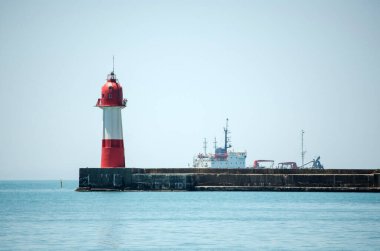 Kızıl deniz feneri berrak gökyüzünün altında, sakin denize bakan bir iskelede duruyor. Arka planda, deniz atmosferini vurgulayan sanayi gemisi yer alıyor. Yumuşak ışıklandırma sakin sahne ve mavi tonları artırır