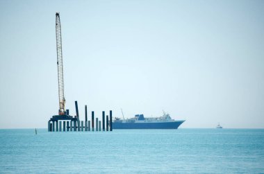 Büyük gemi, kıyı inşaatının yanında sakin mavi denizde yol alır. Uzun vinç platformda dikey direklerle çalışıyor. Temiz gökyüzü huzurlu deniz manzarasını güçlendirir
