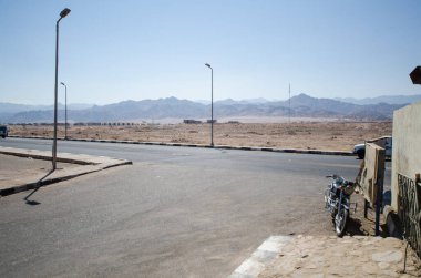 Sakin yol kenarı manzarası, açık gökyüzü, yanına motosiklet park edilmiş. Güneş ışığı altındaki uzak dağlar çöl atmosferini güçlendirir. Sakin ve geniş bir manzara