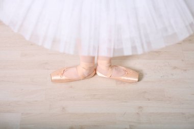 Beyaz tül etekli genç bayan balerin bacakları ahşap zeminde zarif duruyor. Yumuşak ışıklandırma, saten noktalı ayakkabıların içinde dengeli ayakları ön plana çıkarıyor, sakin bir atmosfer yaratıyor.