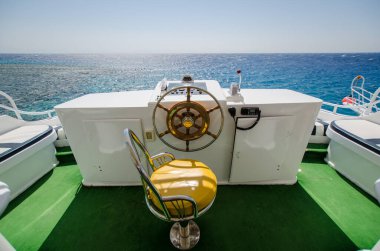 Parlak yeşil zemini ve altın rengi kahverengi koltukları olan lüks bir yat dümeni. Geniş, parlak, açık mavi gökyüzü altında Kızıl Deniz 'e tepeden bakıyor. Güneş ışığı su yüzeyine yansıyor.