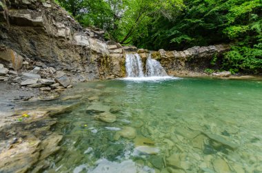 Küçük şelaleler yemyeşil ormanlarla çevrili kristal berraklığında havuza dökülür. Taşlar ve kayalar şeffaf suyun altında görülebilir. Bulutlu aydınlatma huzurlu bir ortam ekler