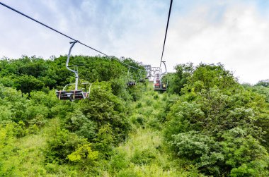 Çelik teleferik kulesi, sık ağaçlarla çevrili yeşil dağ yamacında duruyor. Bulutlu gökyüzü, canlı yeşillikleri vurgulayarak nötr bir zemin oluşturur. Yokuş yukarı bakış açısı yüksekliği ve eğimi vurgular