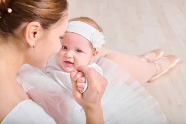Kahverengi saçlı ve açık tenli genç bir kadın bale kıyafeti giymiş gülümseyen bir kız çocuğu tutuyor. Aynı beyaz kıyafetleri giyerek, hafif ahşap zeminde otururlar, sıcaklığı ve bağlantıyı aktarırlar.
