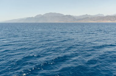 Serene Kızıldeniz suları berrak gökyüzünün altında uzak sıradağlarla buluşuyor. Parlak gün ışığı mavi tonları ve dalgalı yüzeyi vurgular. Huzurlu deniz manzarası