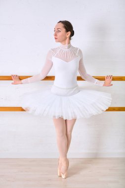 Zarif beyaz etekli genç bayan balerin parlak minimalist stüdyoda zarif bir şekilde duruyor. Yumuşak ışıklandırma, ışık tonlu arka plana karşı sakin ifade ve hassas duruşu vurgular