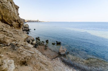 Sakin bir tatil köyü, kayalık kıyı şeridi berrak gökyüzü altında sakin mavi denizle buluşuyor. Sıcak kayaların tonları soğuk su renkleriyle zıtlaşır ve barışçıl bir geri çekilme hissi uyandırır. Yumuşak dalgalar sessizce kıyıya vurur