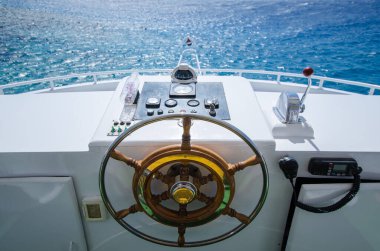 Yat dümeninden parlak güneş ışığı altında çarpıcı Kızıldeniz renkleri görünüyor. Tahta direksiyon ve sakin denizcilik ortamında kayık atmosferini kontrol eder.