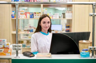 Uzun kahverengi saçlı, genç bir kadın eczacı tezgahın arkasında durup ilaç raflarıyla çevrili parlak ışıklı eczanede gülümsüyor. Profesyonel ve hoş bir ortam.