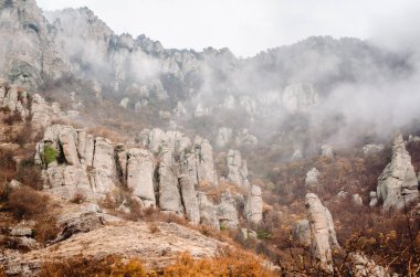 Çarpıcı kaya oluşumları, Kırım Dağları 'ndaki sisli bir zarfın içinden yükseliyor. Doğal manzara sonbahar renkleri ve dramatik jeolojiye sahiptir. Bulutlu gökyüzü ürkütücü, sakin bir atmosfer yaratır.