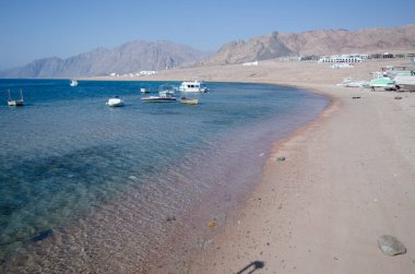 Sakin Kızıldeniz kıyıları, sakin sular, küçük tekneler ve uzak dağlarla. Kumsal, açık mavi gökyüzünün altında davetkar su tonlarıyla uzanır. Doğası gereği büyülenmiş uyum.