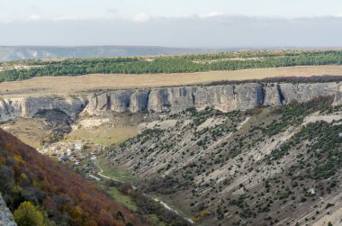 Panoramik manzara engebeli Kırım Vadisi 'ni yakalıyor. Aşağıdaki bulutlu gökyüzünün altındaki küçük köyü saran sarp kayalıklar. Sonbahar renkleri dağınık yeşilliklerle kaplıdır.